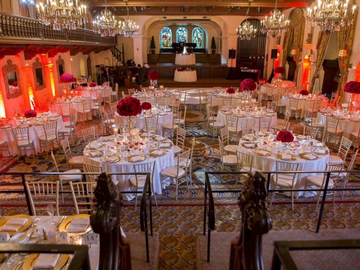 An elegant banquet hall set for a formal event, with chandeliers, lit candelabras, white tablecloths, gold chairs, and red floral decor.