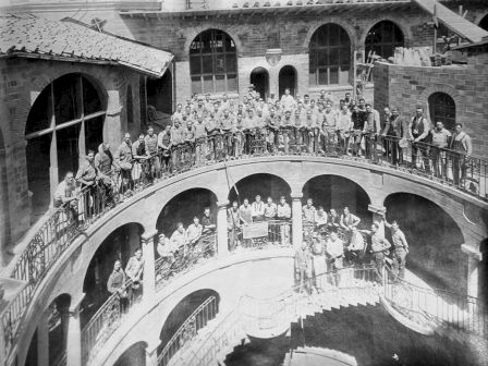 Black-and-white photo of a group posing on a curved, arched walkway in a brick courtyard, with many in uniform and guards.