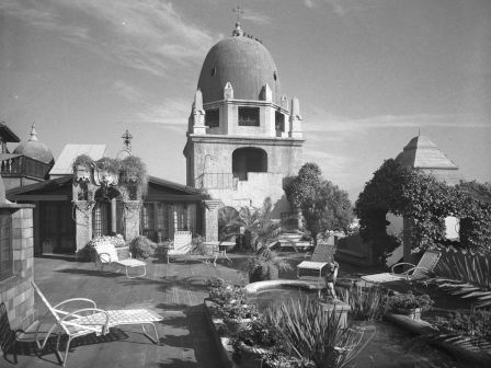 A vintage black-and-white courtyard scene with lounge chairs, lush plants, and a prominent domed building with a small cross on top.