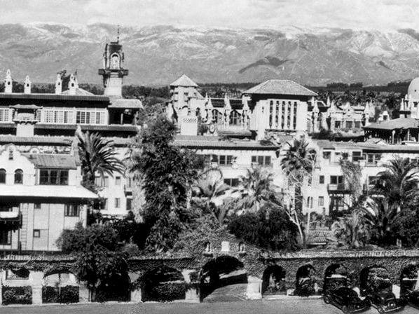 A black-and-white view of a historic mission complex with church towers, gardens, parked cars, and mountains in the background.