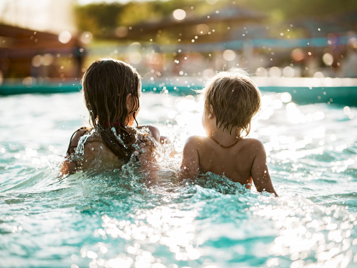 Two children splash in a sunny pool, seen from behind as they laugh and play in sparkling water.