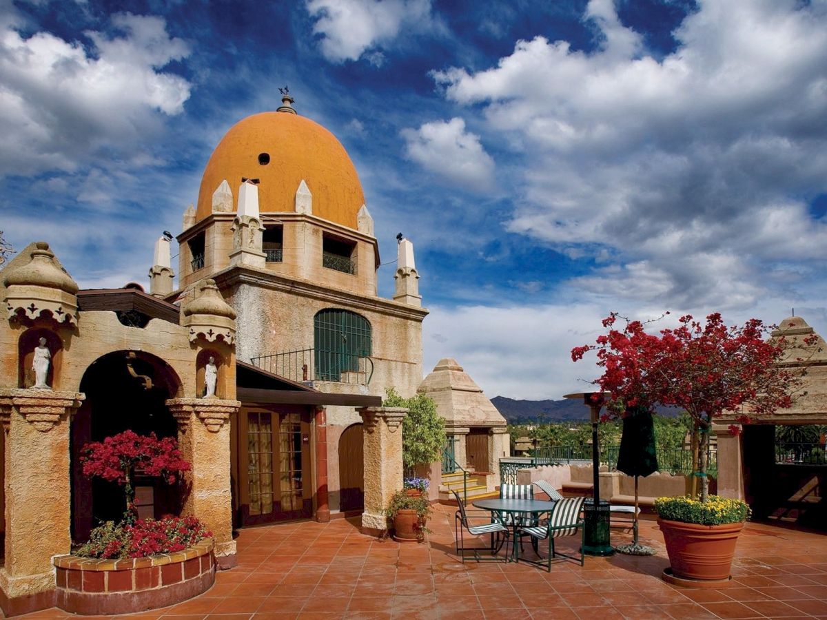 A sunny courtyard with an orange-domed church, arches, patio seating, and blooming red flowers under a blue sky.