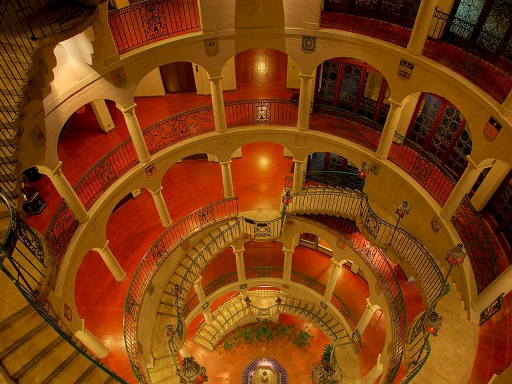 An ornate interior with a sweeping spiral staircase, arched balconies, warm red floors, and decorative lamps and statues.