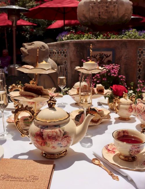 An ornate outdoor afternoon tea table with a floral teapot, cups, silverware, and a flowered garden backdrop under red umbrellas.