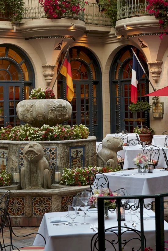An elegant outdoor courtyard set for dining, with bunting flags, flowers, and stone statues in an arched garden.