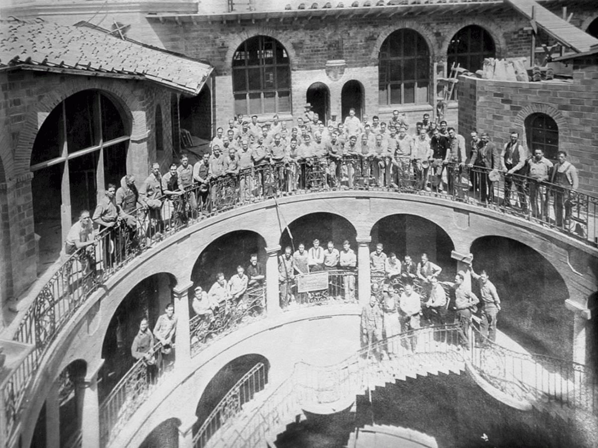 Black-and-white photo of a brass band lined up inside a stone courtyard with arched bridges and onlookers.