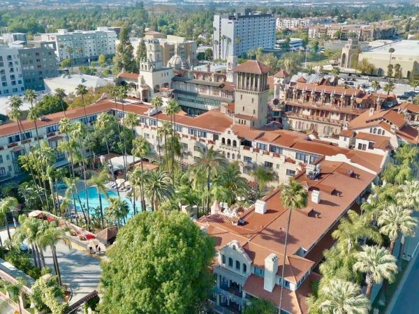 Aerial view of a sunny resort with Mediterranean-style buildings, red-tiled roofs, palm trees, and a blue pool courtyard.